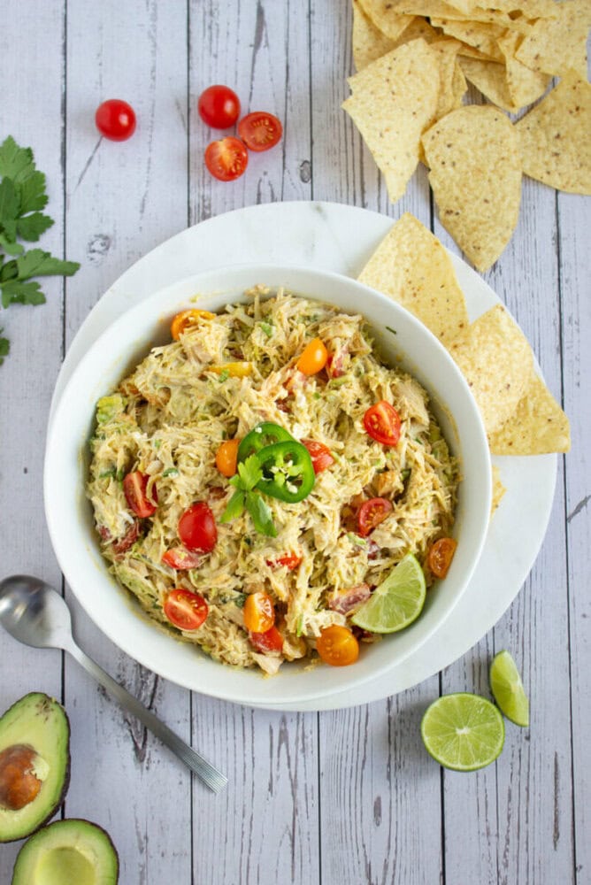 A bowl of shredded chicken salad garnished with cherry tomatoes, jalapeño slices, cilantro, and lime, served with tortilla chips on a white wooden surface.