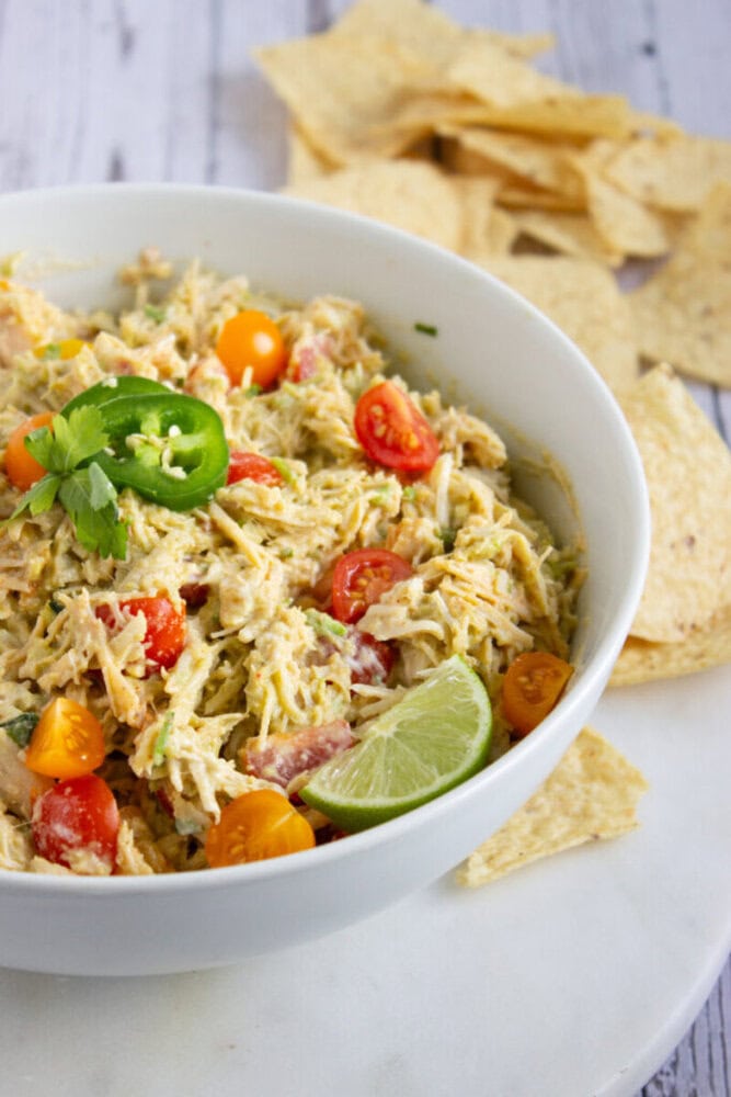 A white bowl filled with creamy shredded chicken salad, cherry tomatoes, a lime wedge, jalapeño slice, and cilantro, with tortilla chips on the side.