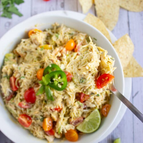 A bowl of shredded chicken dip with sliced cherry tomatoes, jalapeños, cilantro, and lime, served with tortilla chips on a wooden surface.