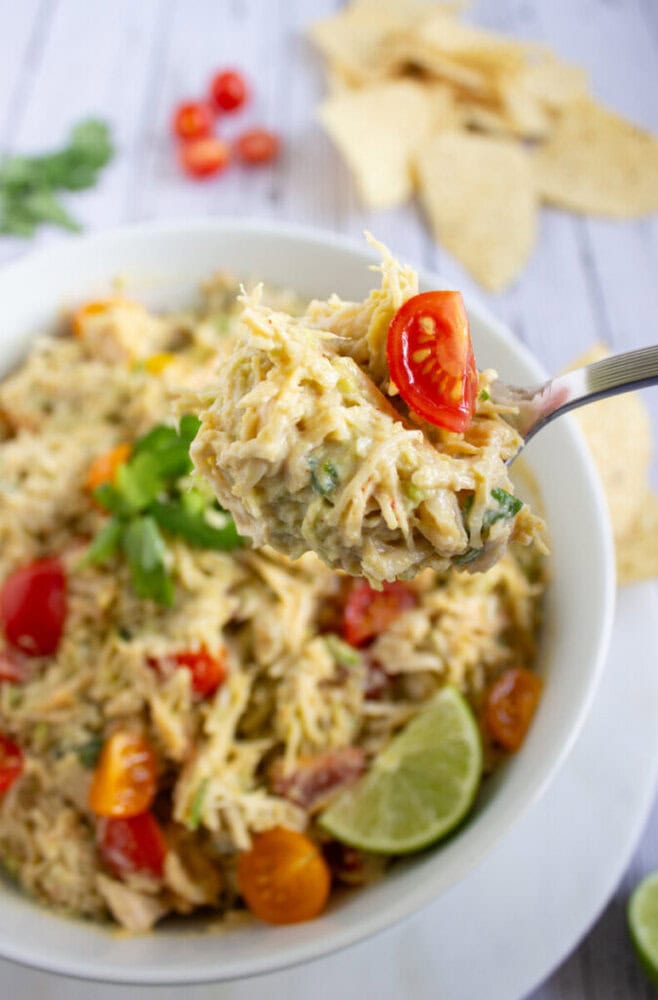 A spoonful of shredded chicken dip with tomatoes and herbs is held above a bowl, with tortilla chips and lime visible in the background.