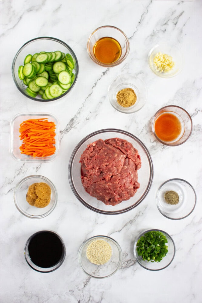 A flat lay of various ingredients in glass bowls, including ground meat, sliced cucumbers, carrots, sauces, seasonings, garlic, green onions, and sesame seeds, on a marble surface.