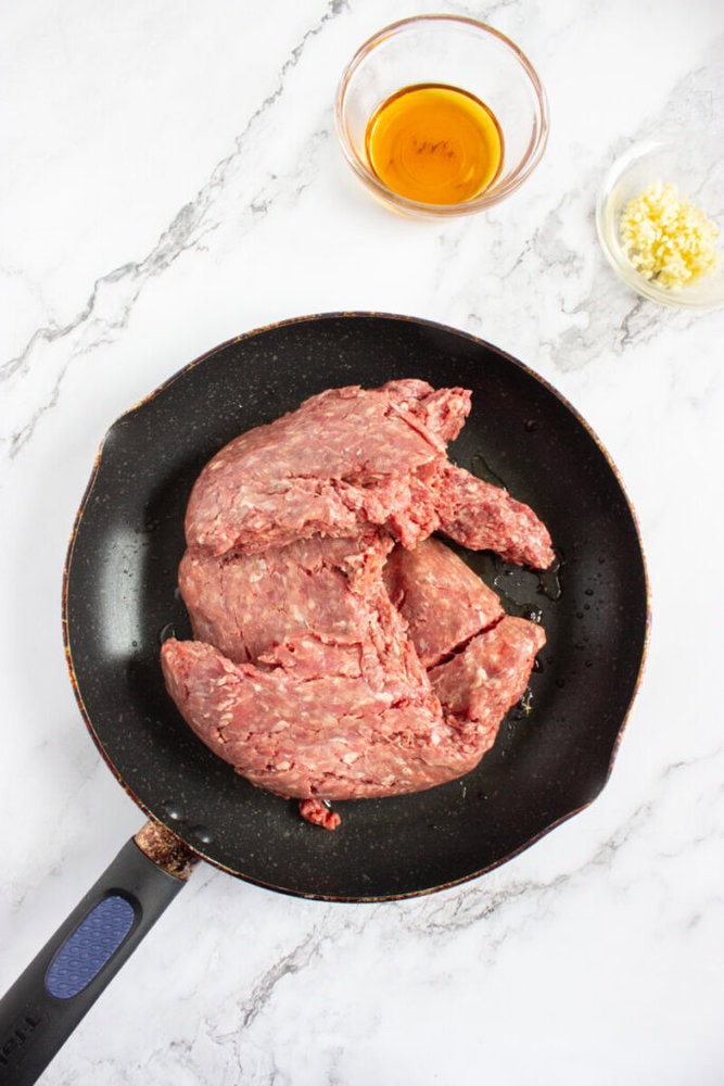 Raw ground meat in a nonstick frying pan, with a small bowl of minced garlic and a bowl of oil on a white marble surface.