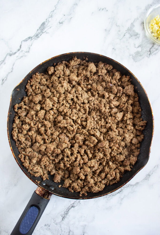 A skillet filled with cooked ground beef sits on a marble countertop, with a small glass bowl of minced garlic nearby.