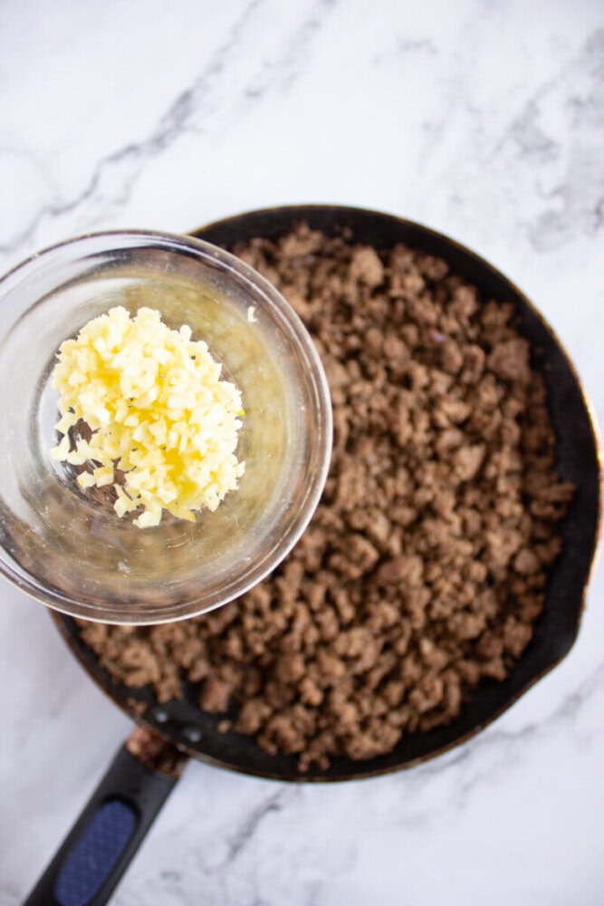 A glass bowl of minced garlic is held over a skillet filled with cooked ground beef on a marble countertop.