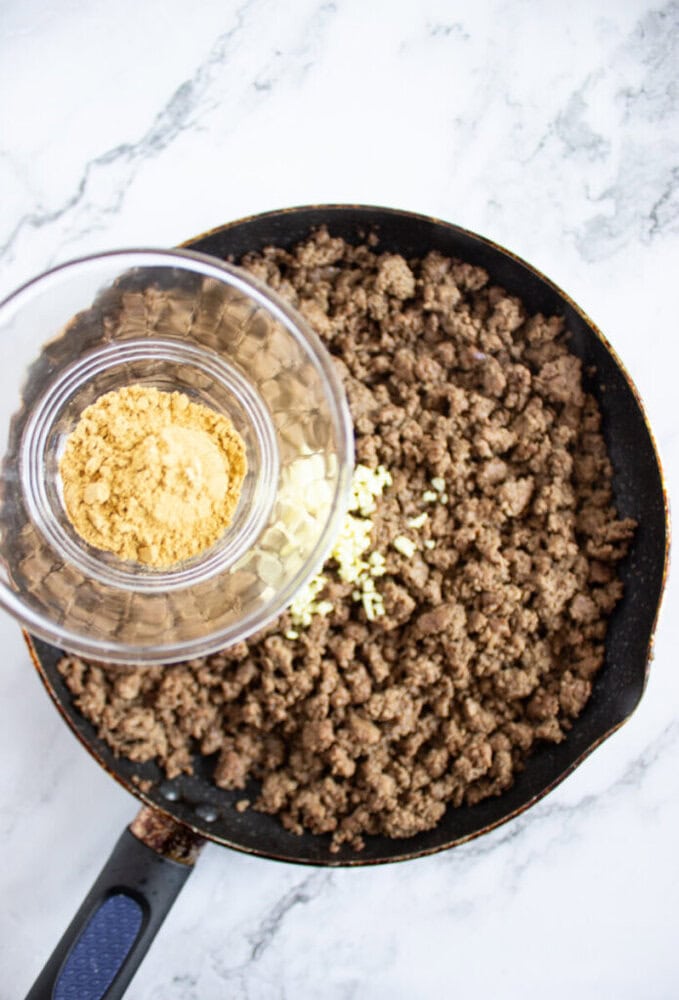 Cooked ground beef in a frying pan on a marble surface, with a glass bowl of seasonings and minced garlic being held above the pan.