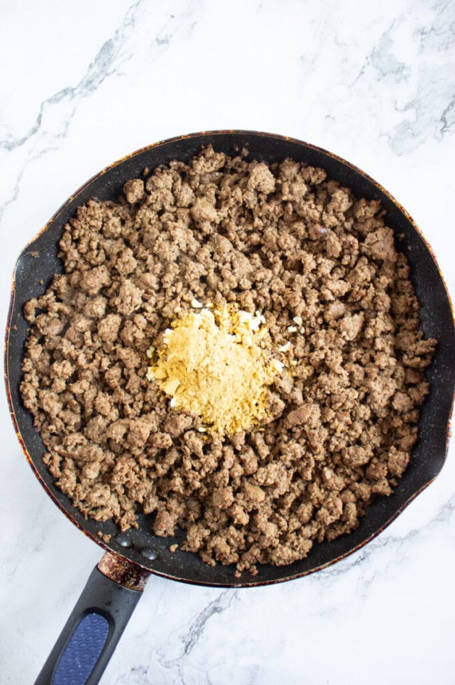 Cooked ground beef in a skillet with a mound of seasoning powder placed in the center, on a light marble surface.