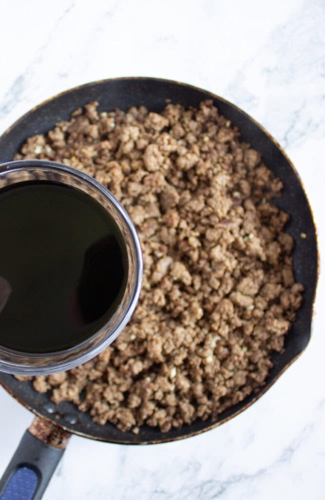 A skillet filled with cooked ground beef next to a measuring cup containing a dark liquid, on a marble surface.