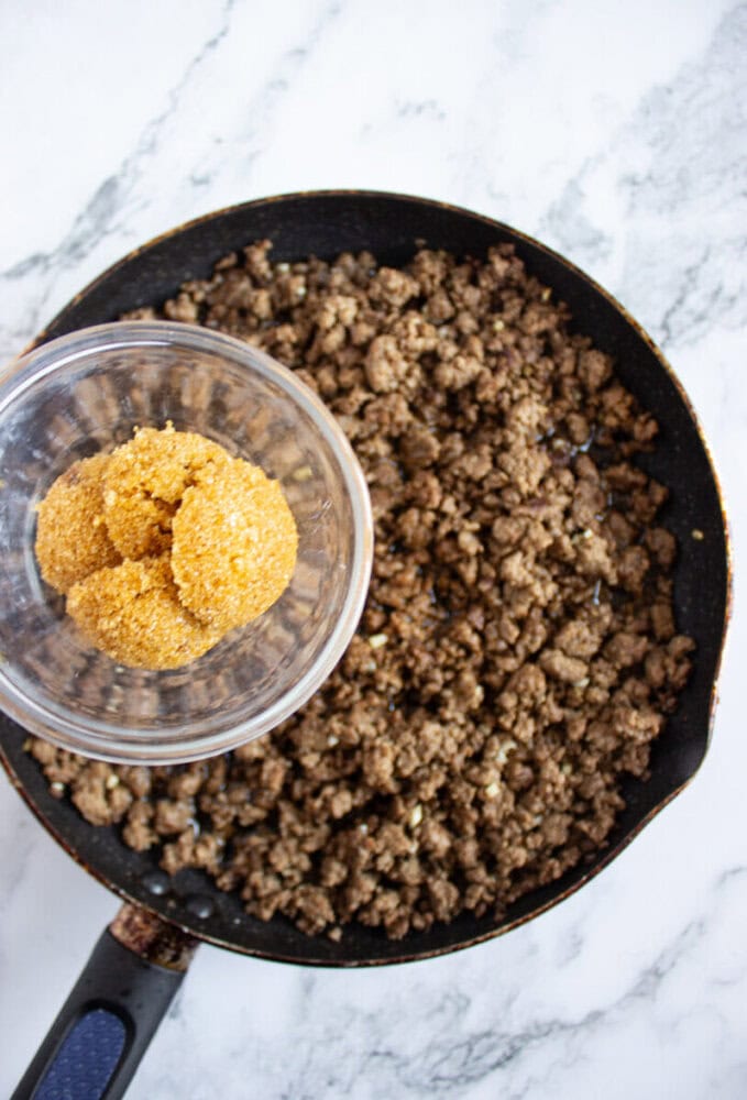 Cooked ground beef in a skillet with a glass bowl of taco seasoning being held above it, on a marble countertop.