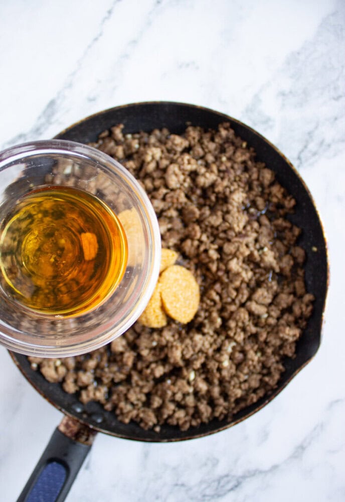 Ground meat cooking in a skillet with two piles of seasoning; a hand holds a glass measuring cup with a yellow-brown liquid above the pan.