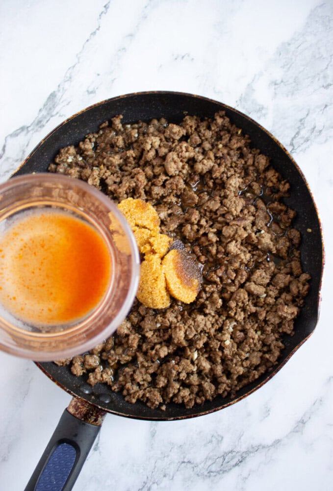 Ground beef is cooking in a skillet with seasoning and orange liquid being poured in from a measuring cup, all on a marble countertop.