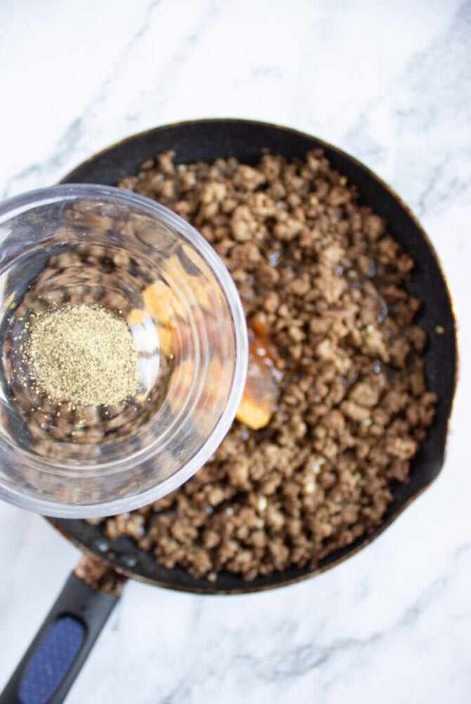 Ground beef cooking in a skillet, with a glass container of black pepper being held above, ready to be added.