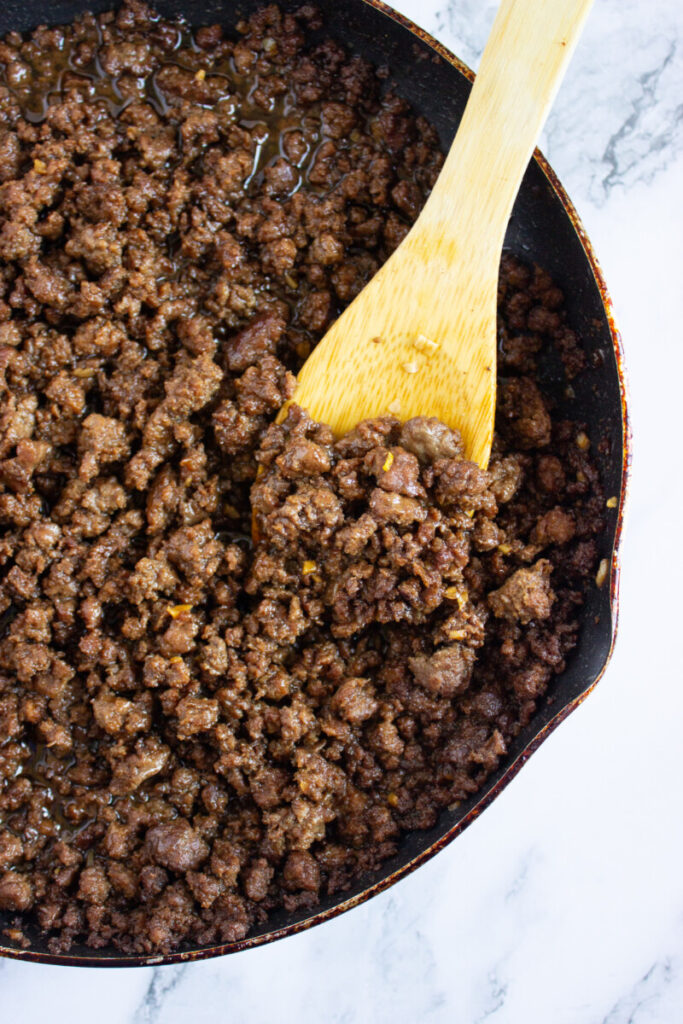 Cooked ground beef with visible seasoning in a skillet, being stirred with a wooden spoon on a marble surface.