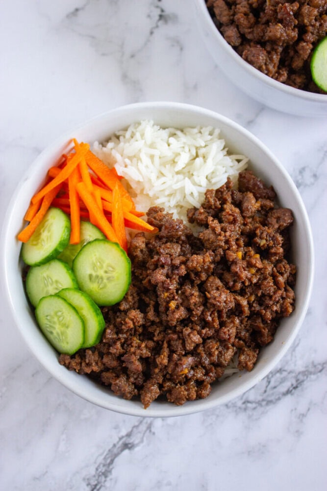 A bowl containing ground beef, white rice, sliced cucumbers, and julienned carrots on a marble surface.