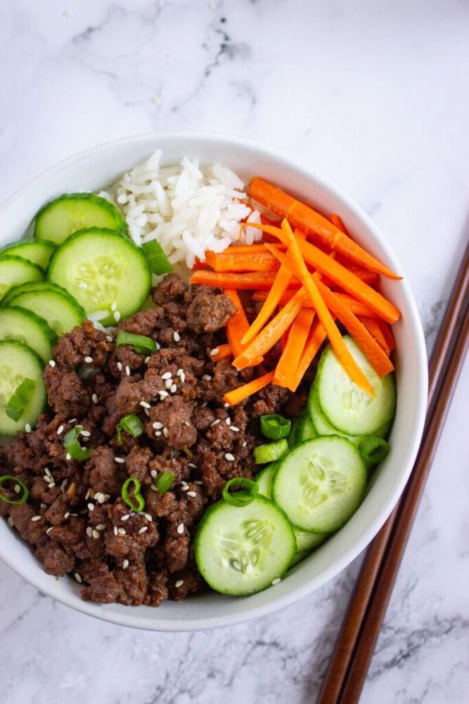 A bowl containing ground beef with sesame seeds, white rice, sliced cucumbers, and julienned carrots on a marble surface with chopsticks beside it.