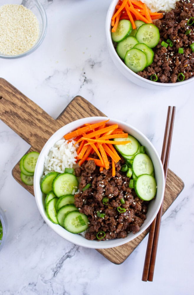 A bowl with ground beef, white rice, sliced cucumbers, julienned carrots, and green onions, with chopsticks resting on the side. Another similar bowl and a bowl of sesame seeds are nearby.