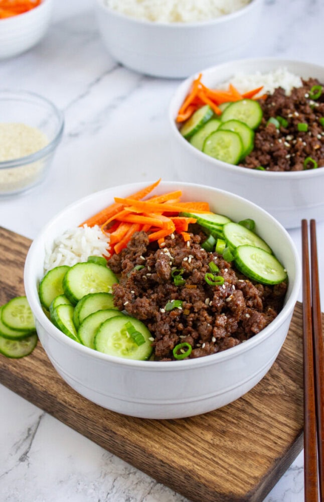 A white bowl filled with white rice, seasoned ground beef, sliced cucumbers, shredded carrots, and topped with green onions and sesame seeds. Chopsticks rest nearby.