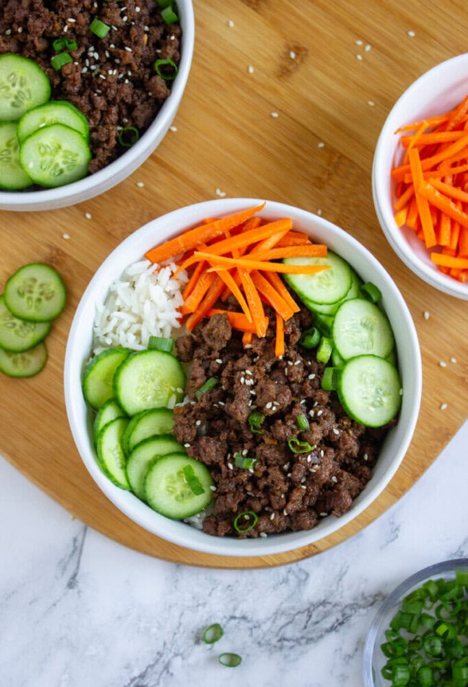 A bowl containing white rice, sliced cucumbers, shredded carrots, and seasoned ground beef, garnished with green onions and sesame seeds, on a wooden board.