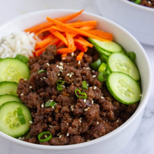 A white bowl filled with ground beef, sliced cucumbers, carrot sticks, rice, and garnished with sesame seeds and green onions.