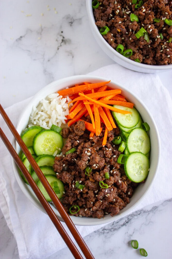 A bowl with cooked ground beef, sliced cucumbers, carrot sticks, white rice, and chopped green onions, garnished with sesame seeds. Chopsticks rest next to the bowl.