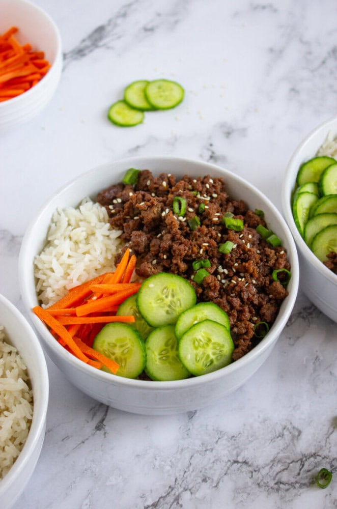 A bowl filled with white rice, sliced cucumbers, shredded carrots, and ground beef topped with green onions and sesame seeds.