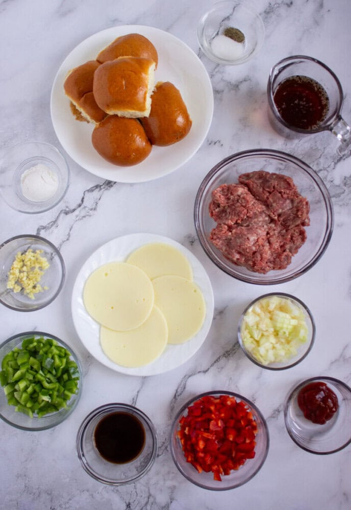 Ingredients for sliders arranged on a marble surface, including buns, ground meat, cheese slices, chopped peppers, onion, garlic, sauces, and seasonings in separate bowls.