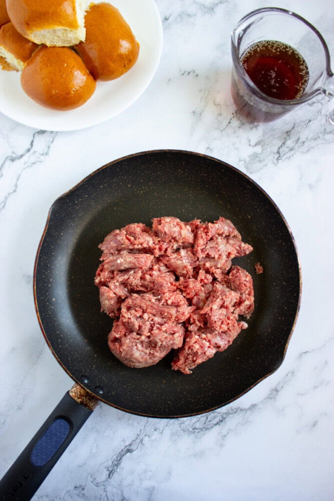 Raw ground beef in a frying pan, with a plate of slider buns and a measuring cup of liquid on a marble countertop.