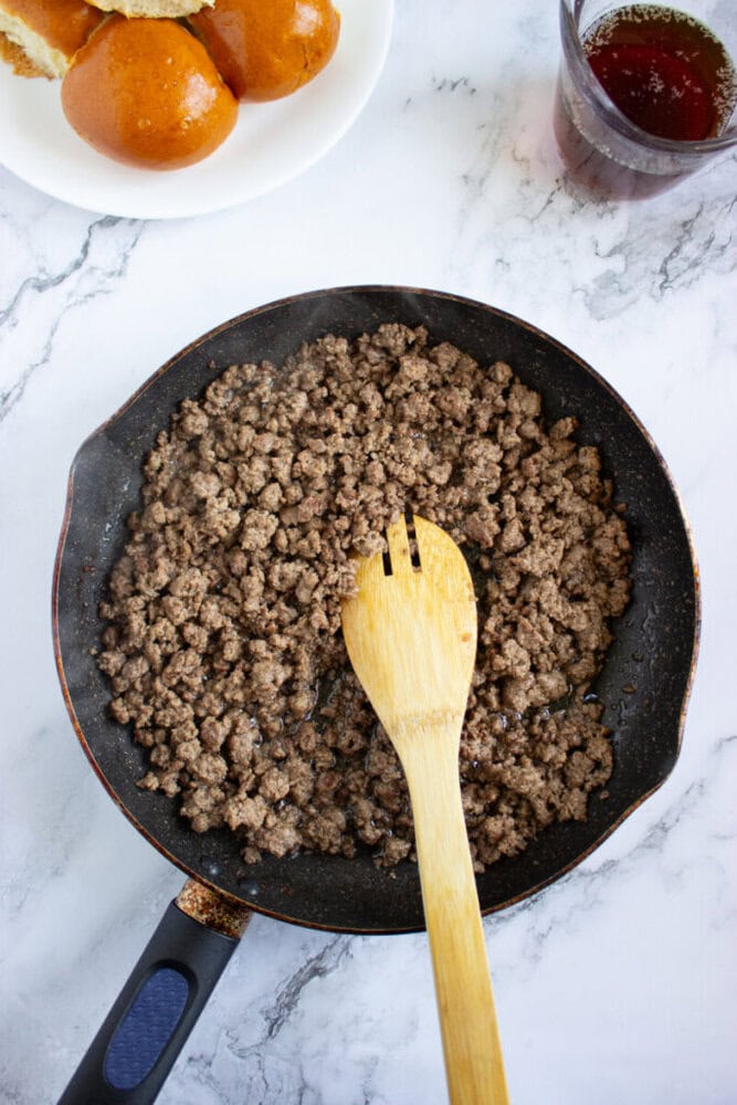 A frying pan with cooked ground beef and a wooden spoon sits on a marble surface, with a plate of buns and a glass of liquid nearby.