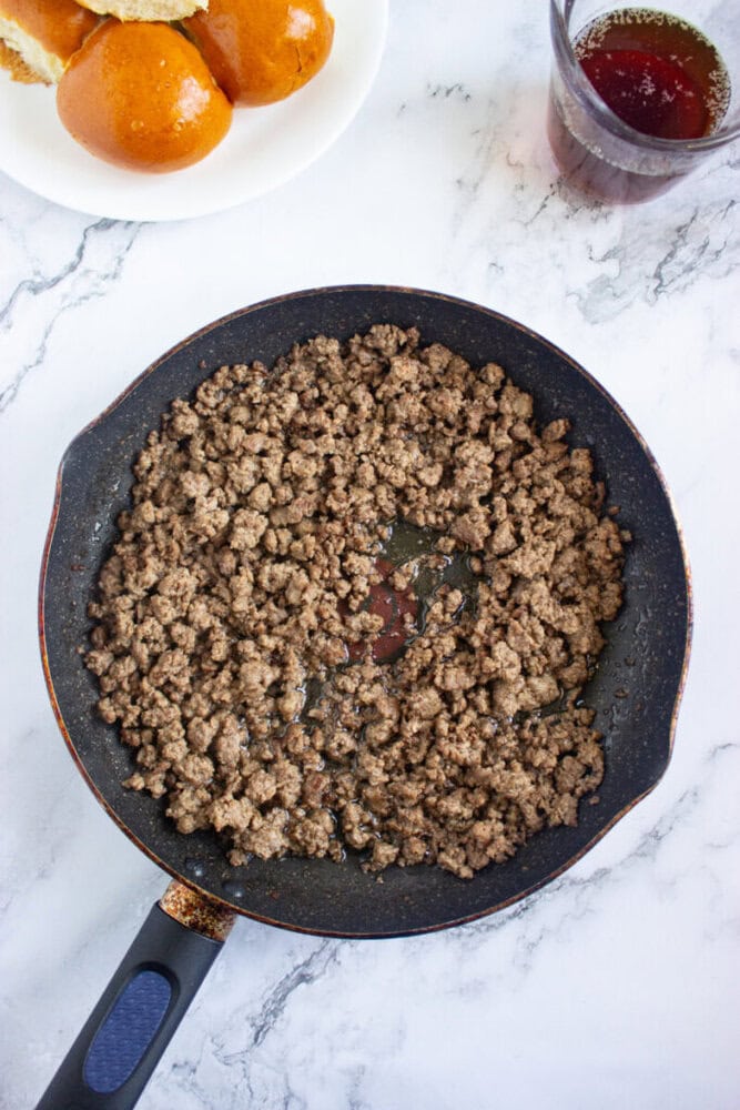 A skillet filled with cooked ground beef sits on a marble surface, with a plate of brioche buns and a glass of drink nearby.