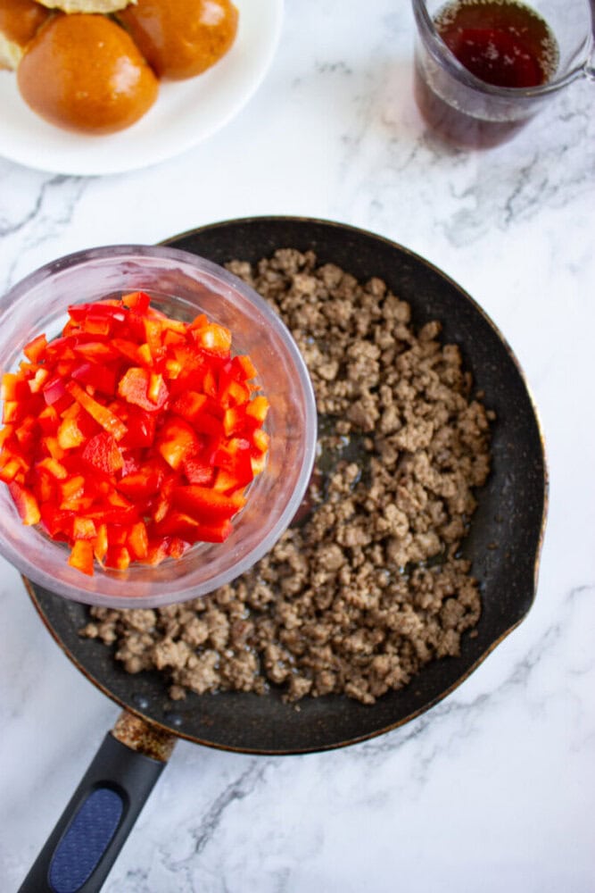 A skillet with cooked ground beef sits on a marble surface. A bowl of chopped red bell peppers is held above the skillet, ready to be added. Nearby are burger buns and a measuring cup with liquid.