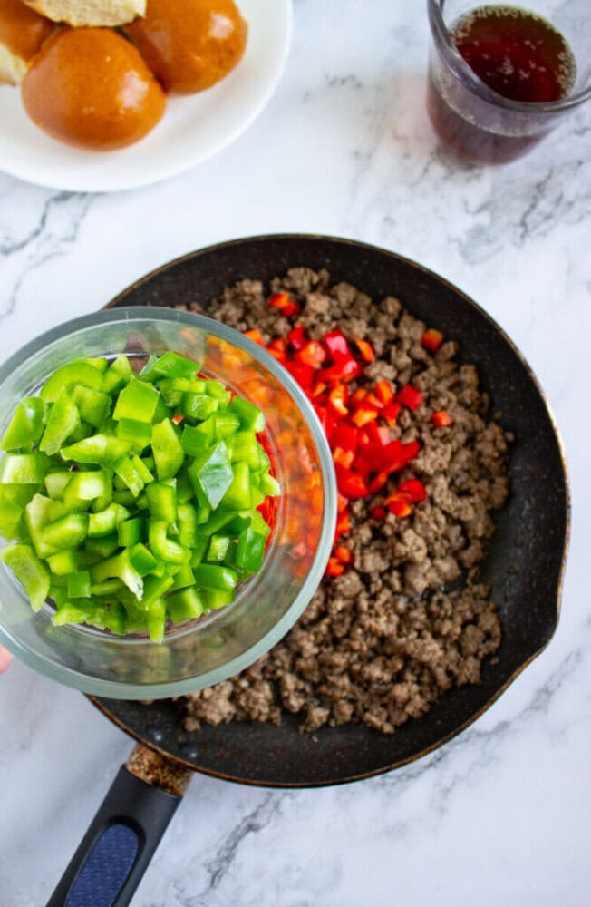 A hand holds a bowl of chopped green bell peppers above a skillet with cooked ground beef and chopped red peppers on a marble countertop. Buns and a drink are in the background.