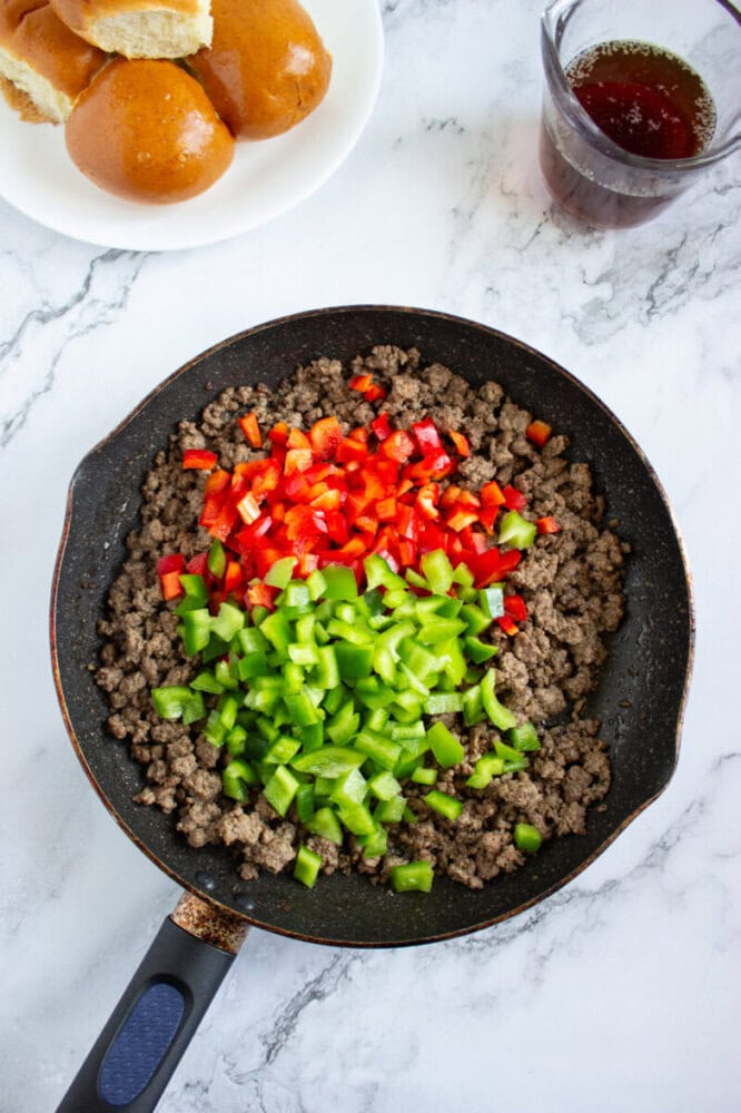 A skillet with cooked ground beef topped with chopped red and green bell peppers, next to a plate of bread rolls and a cup of dark liquid on a marble surface.