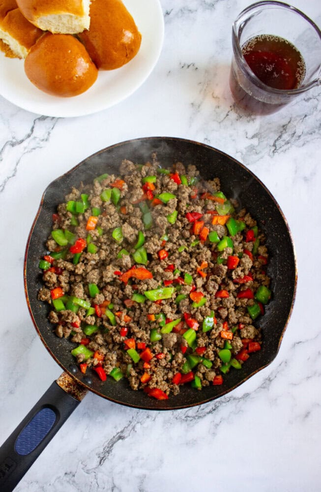 A skillet with cooked ground beef, green and red bell peppers. Plate with bread rolls and a glass of dark liquid are on a marble countertop.