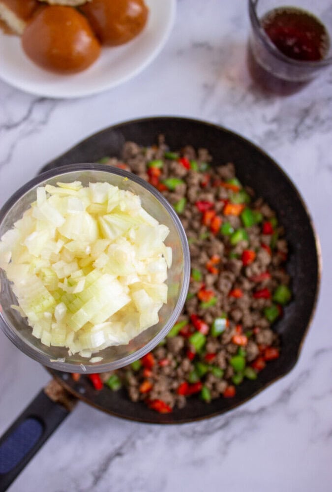 A bowl of chopped onions is held above a skillet with cooked ground beef and diced red and green bell peppers. Buns and a drink are in the background.