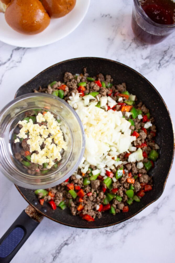 A skillet with cooked ground beef, chopped green and red bell peppers, and chopped onions, with a bowl of minced garlic being added. A plate of buns and a drink are in the background.