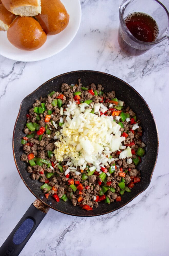 A frying pan with cooked ground beef, chopped red and green bell peppers, topped with diced onions and minced garlic on a marble countertop. Bread rolls and a glass of sauce are nearby.