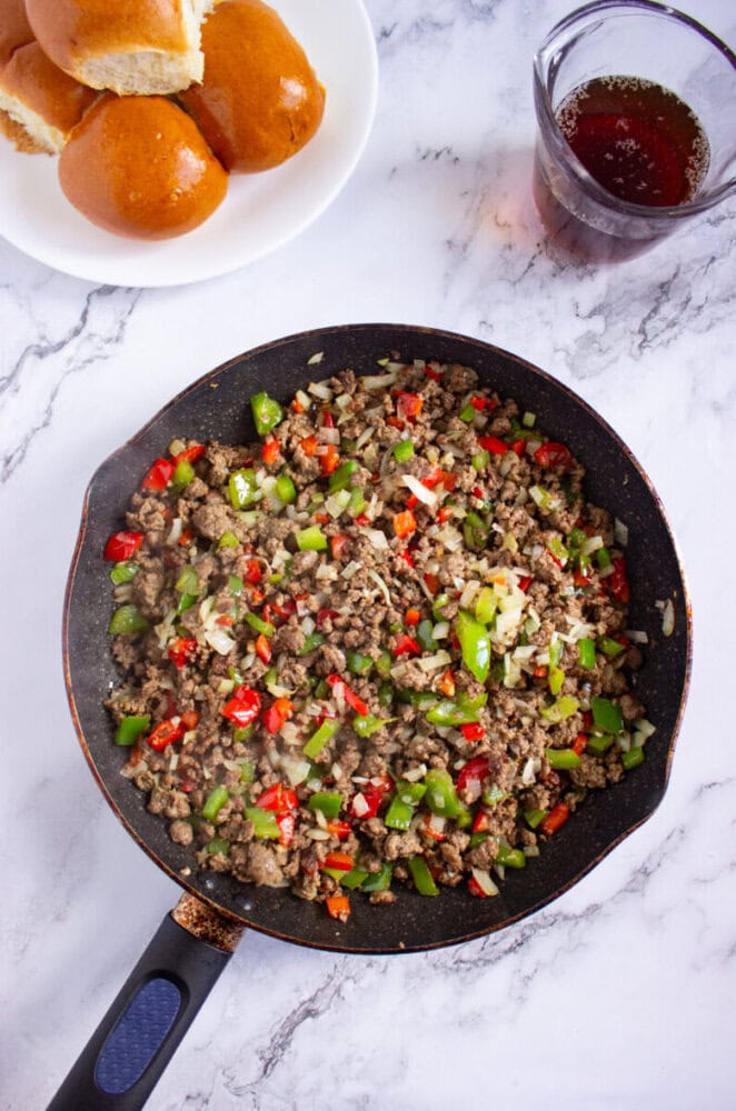 A skillet filled with cooked ground beef, chopped green and red bell peppers, and onions sits on a marble surface next to a plate of bread rolls and a glass of dark liquid.