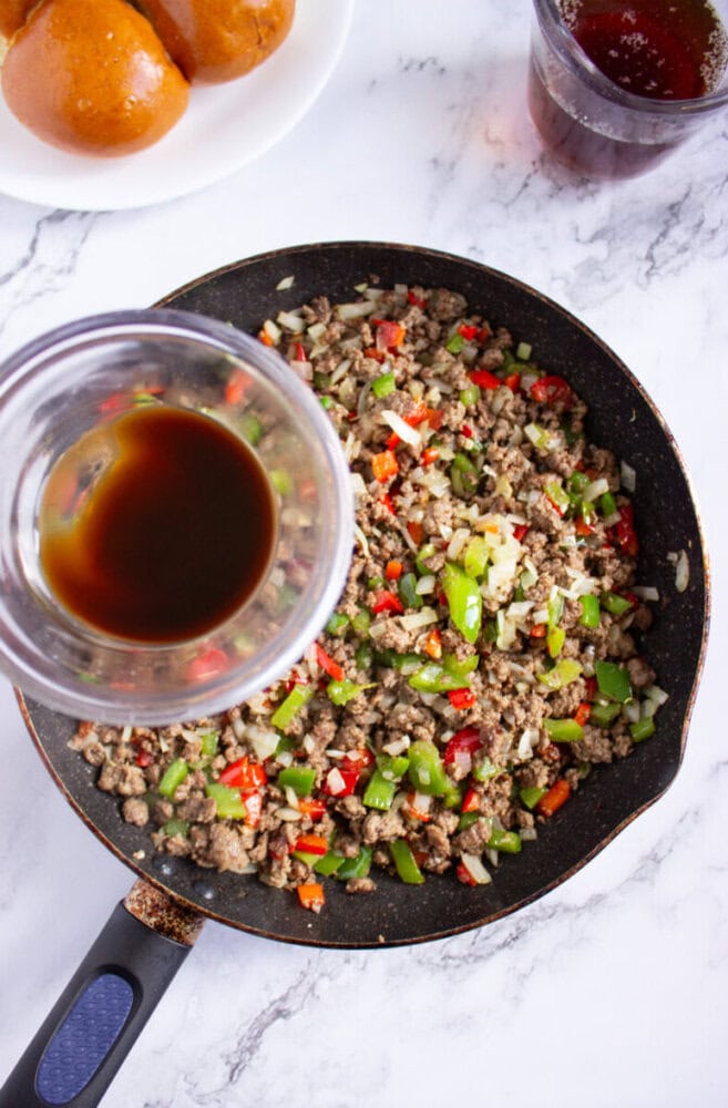 A skillet with ground beef, chopped bell peppers, and onions on a marble surface. A glass with dark liquid is being poured into the skillet. A plate of buns and a drink are nearby.