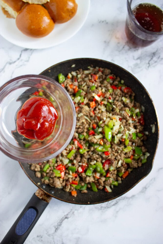 A skillet with cooked ground beef, green and red bell peppers, and onions is on a marble surface. A hand holds a bowl of ketchup above the skillet. Rolls and a drink are in the background.