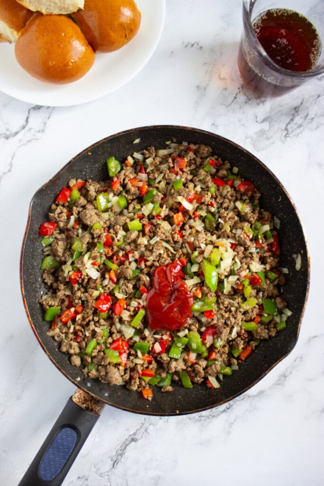 A frying pan contains cooked ground beef mixed with diced red and green bell peppers and onions, topped with ketchup. A plate of bread rolls and a glass of dark liquid are nearby.