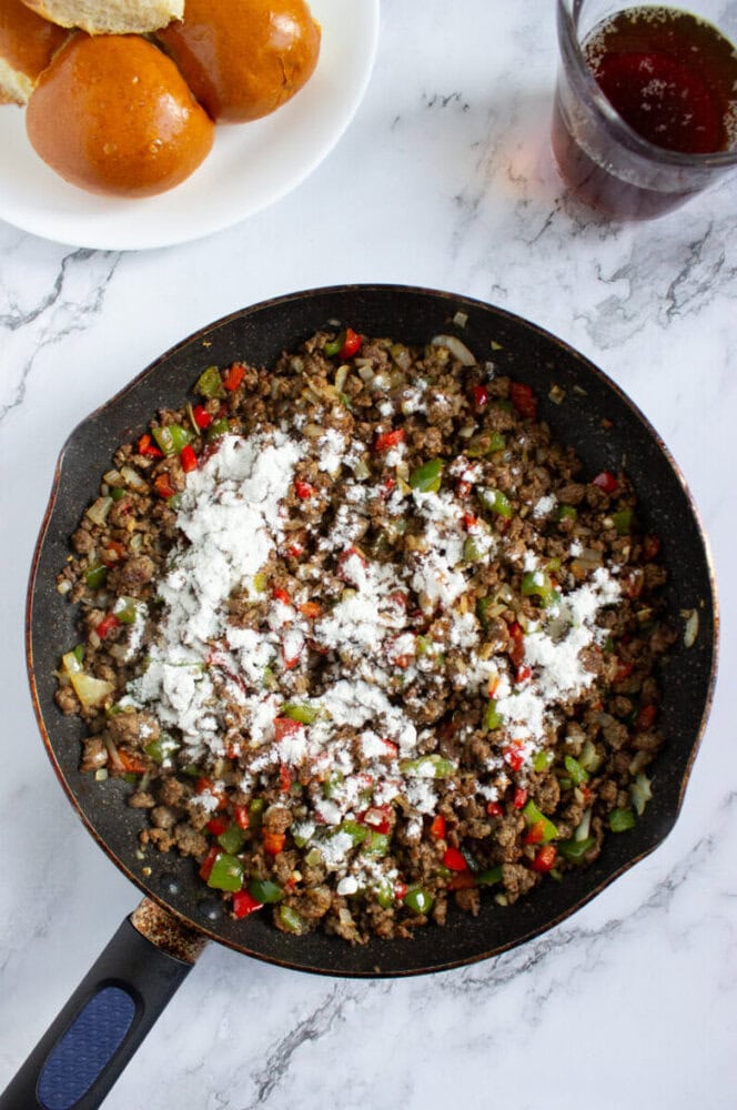 A skillet filled with browned ground beef, diced green and red bell peppers, onions, and a layer of white flour on top. A plate of buns and a glass of dark liquid are nearby.