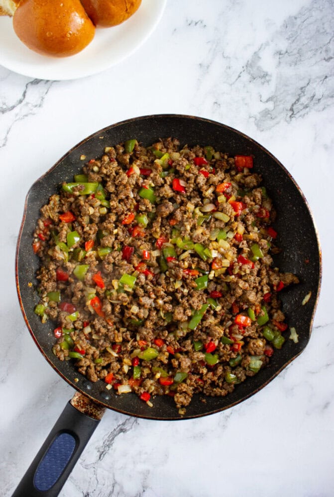 A skillet filled with cooked ground beef, chopped green and red bell peppers, and onions sits on a marble countertop with a plate of sandwich buns nearby.