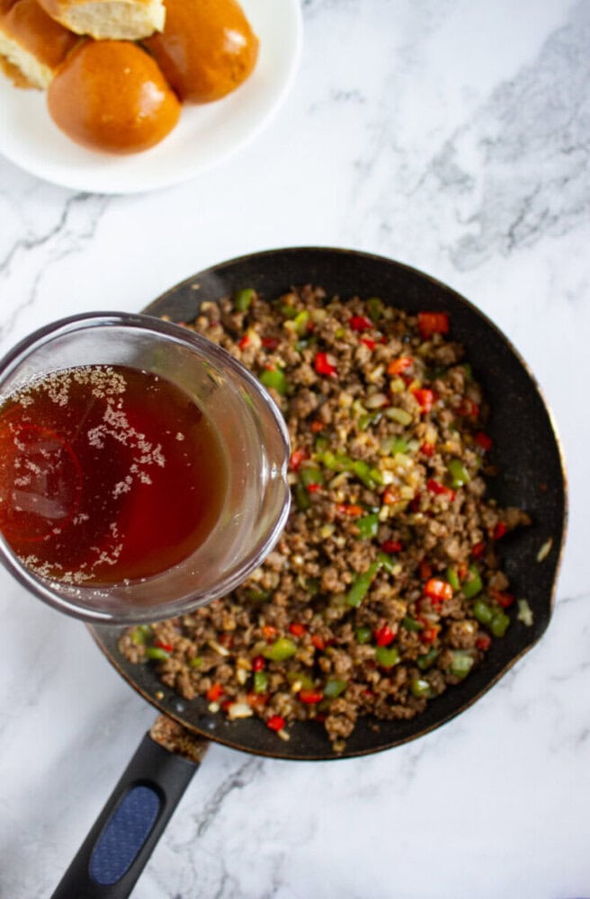 A glass of brown liquid is held above a skillet filled with cooked ground beef and chopped vegetables; a plate of bread rolls is in the background.