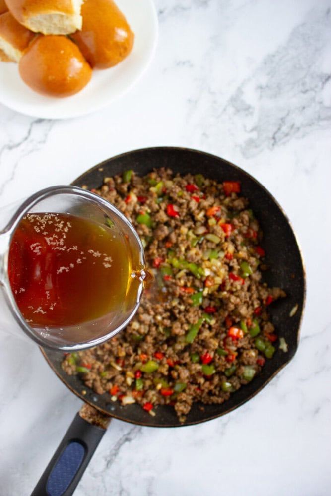 A measuring cup with brown liquid is being poured into a skillet of ground beef and chopped vegetables, with a plate of bread rolls nearby.