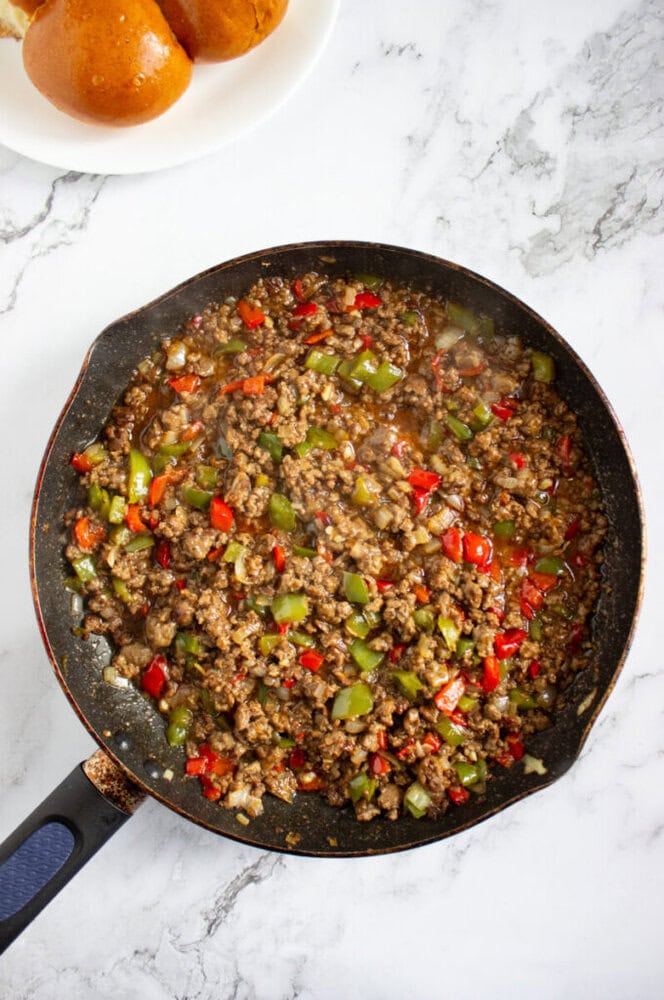 A skillet filled with cooked ground beef, diced green and red bell peppers, and onions sits on a marble countertop; a plate with two buns is in the corner.