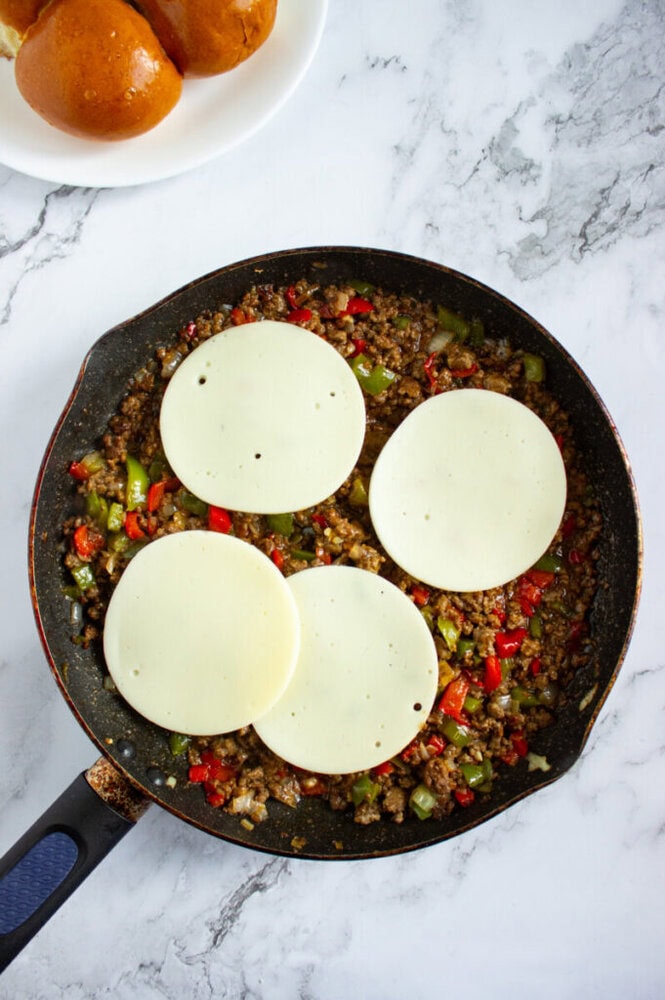 A skillet filled with cooked ground meat, diced vegetables, and four slices of cheese on top, with a plate of sandwich buns in the background.