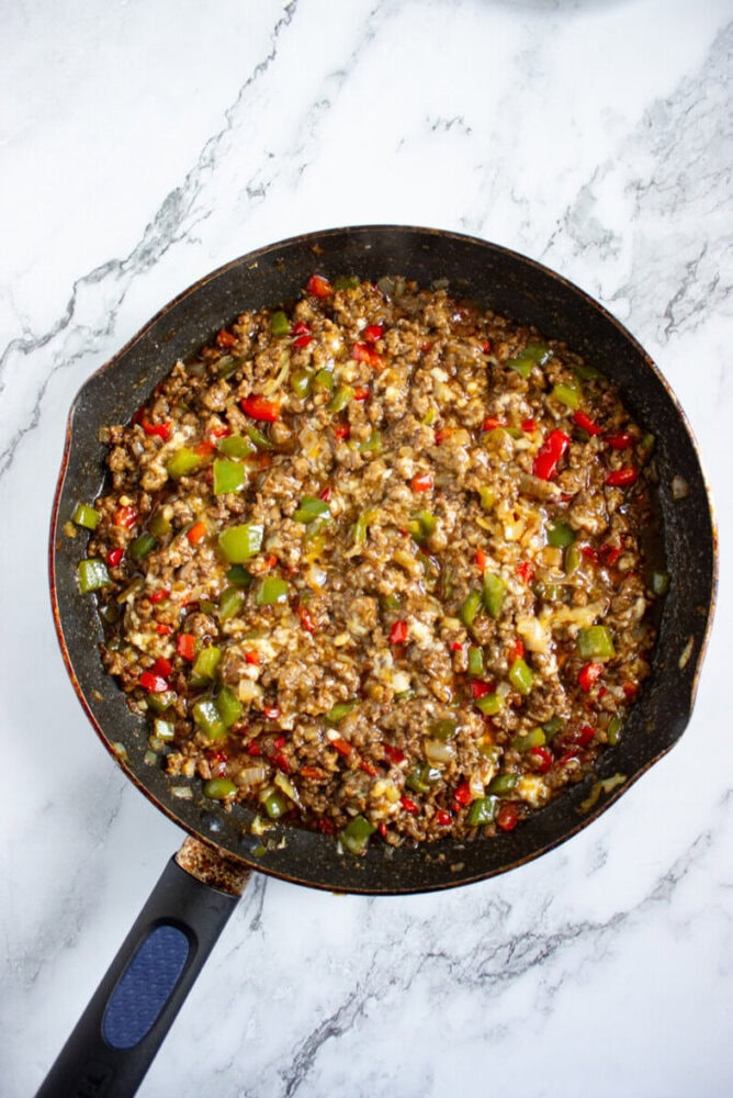 A skillet filled with a cooked mixture of ground meat, diced green and red bell peppers, and onions on a marble countertop.