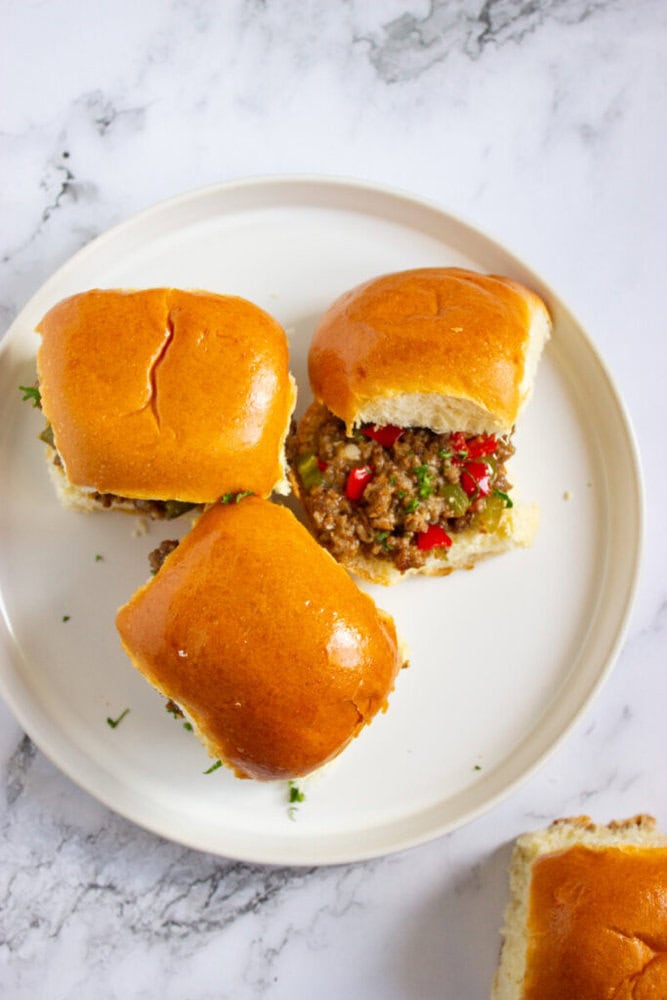 Three beef sliders with glossy buns are arranged on a white plate, one slider showing a ground beef filling with chopped red and green peppers.