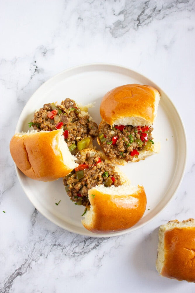 Three sloppy joe sandwiches with ground meat and diced peppers filling are on a white plate, with one additional sandwich on the marble surface beside the plate.
