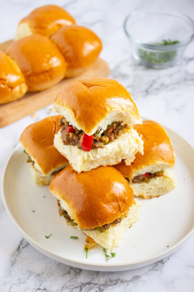 Three filled sliders are stacked on a white plate, with a cutting board of plain baked rolls and a glass bowl in the background on a marble surface.