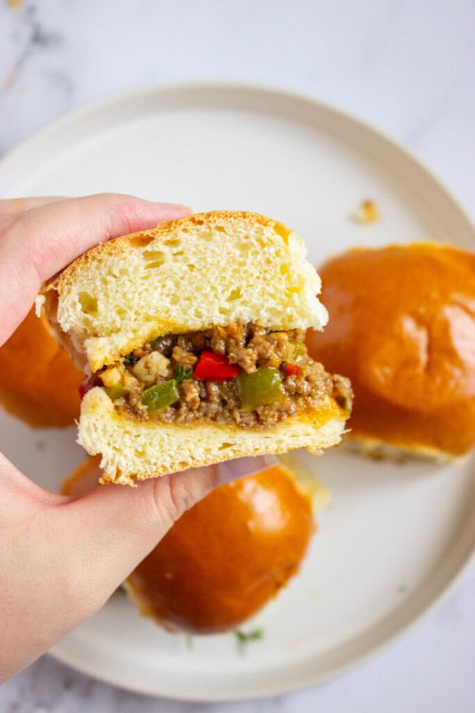 A hand holding half of a sandwich with seasoned ground meat and diced peppers, with two whole sandwiches on a white plate in the background.